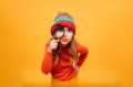 Joyful Young girl in sweater and hat looking at the camera with magnifier over orange background